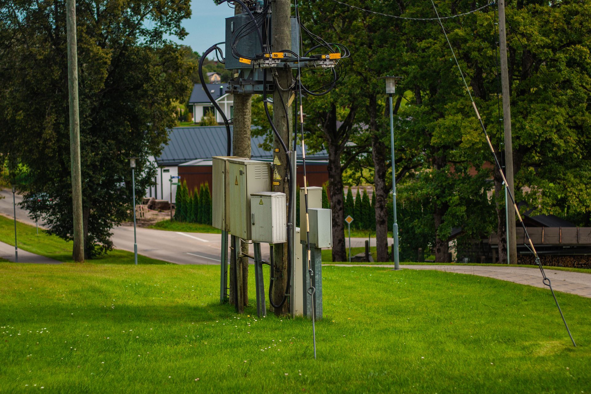 Utility pole with boxes on a grassy area, a road, and buildings with trees in the background.