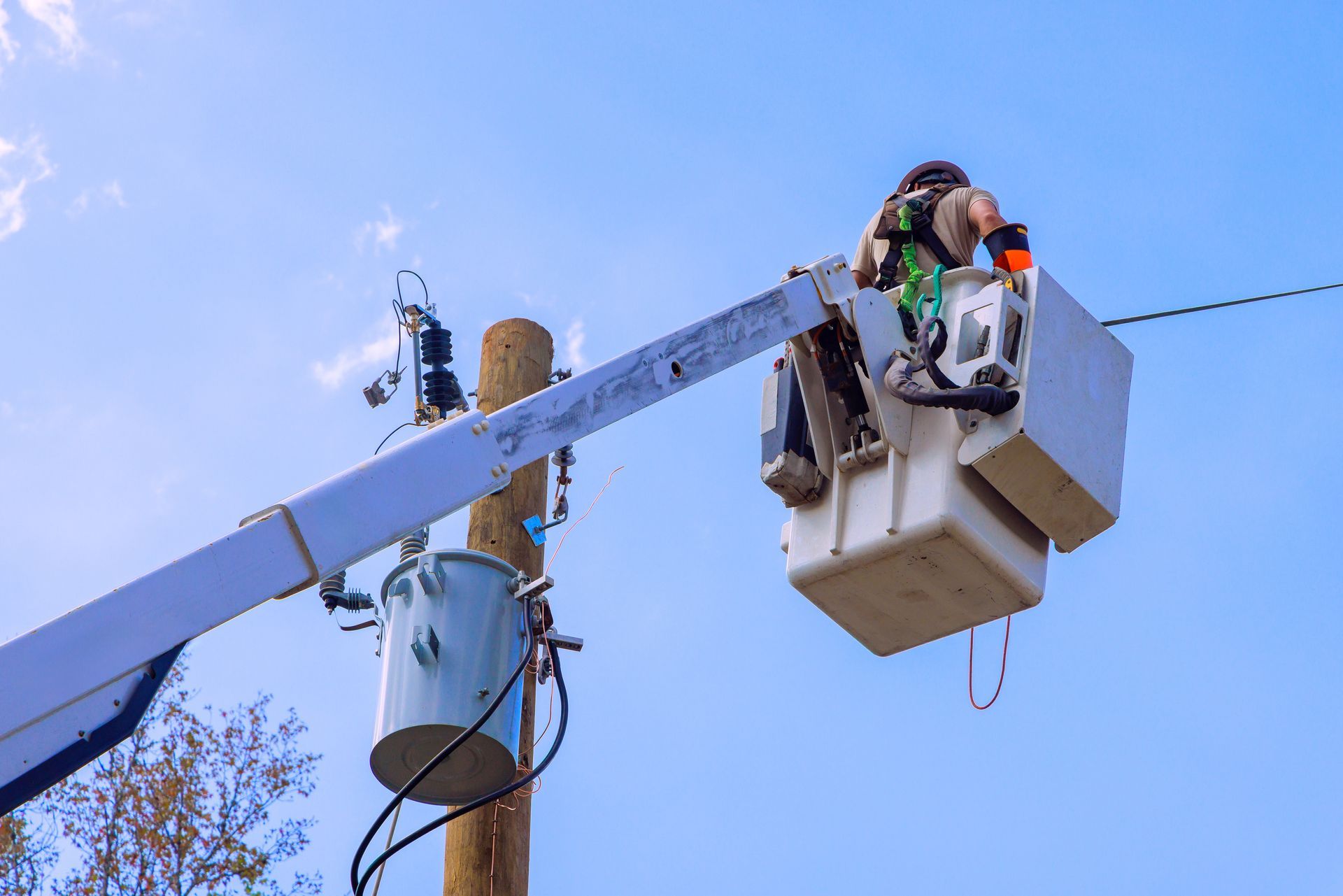 Lineman in lift bucket working on power lines attached to a utility pole against a blue sky.