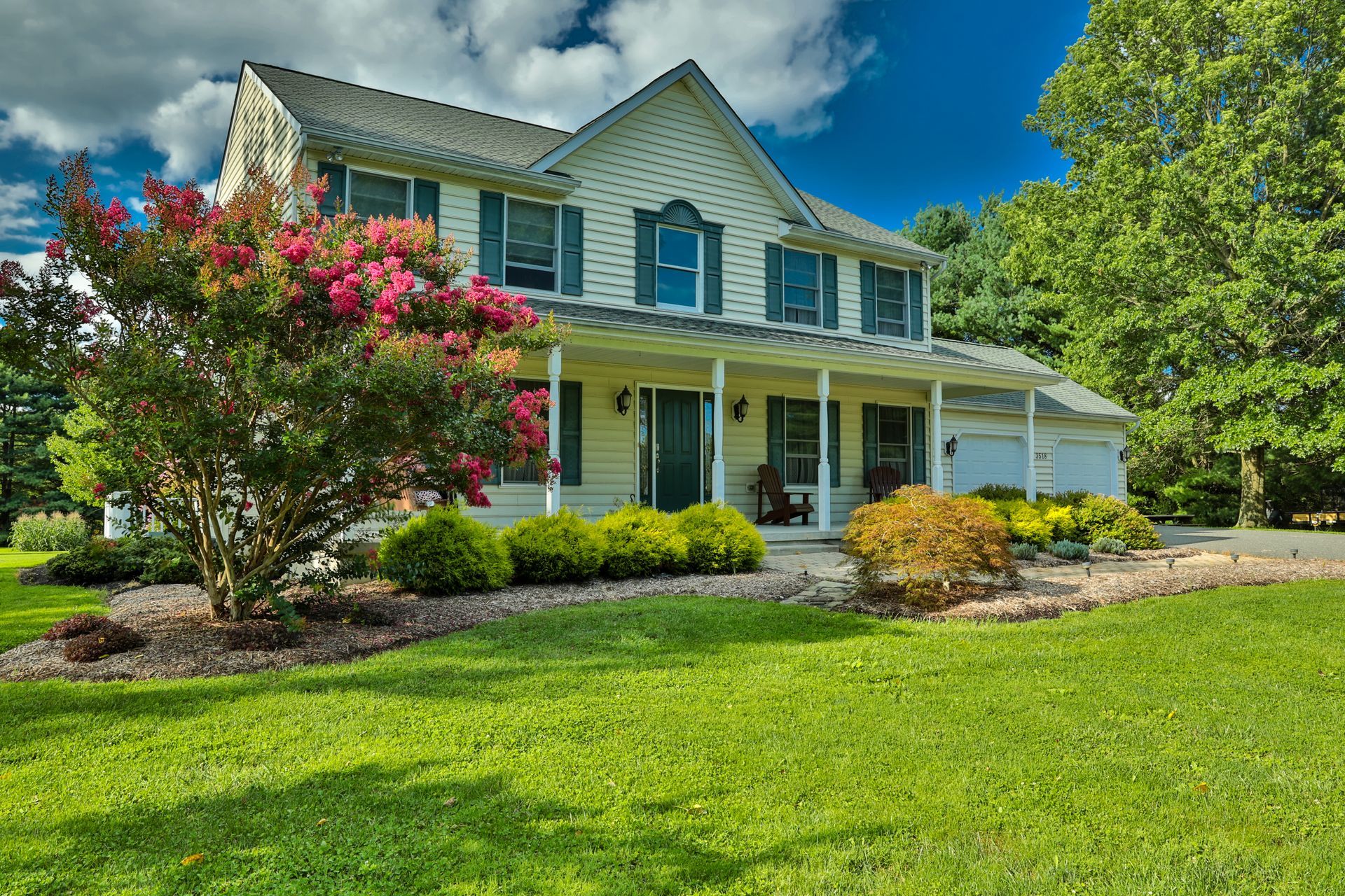 Two-story beige house with green lawn, covered porch, blue shutters, and blooming pink tree.