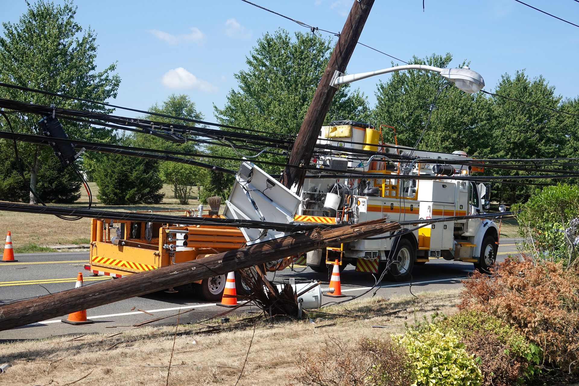 Utility truck near fallen power pole, with lines tangled. Orange cones and trees in background.