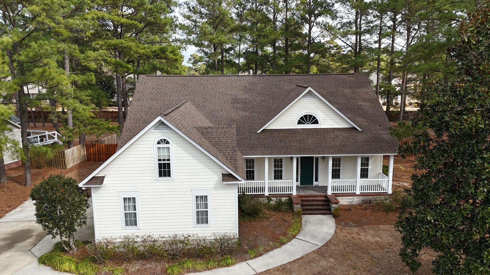 An aerial view of a white house with a new asphalt shingle roof surrounded by trees