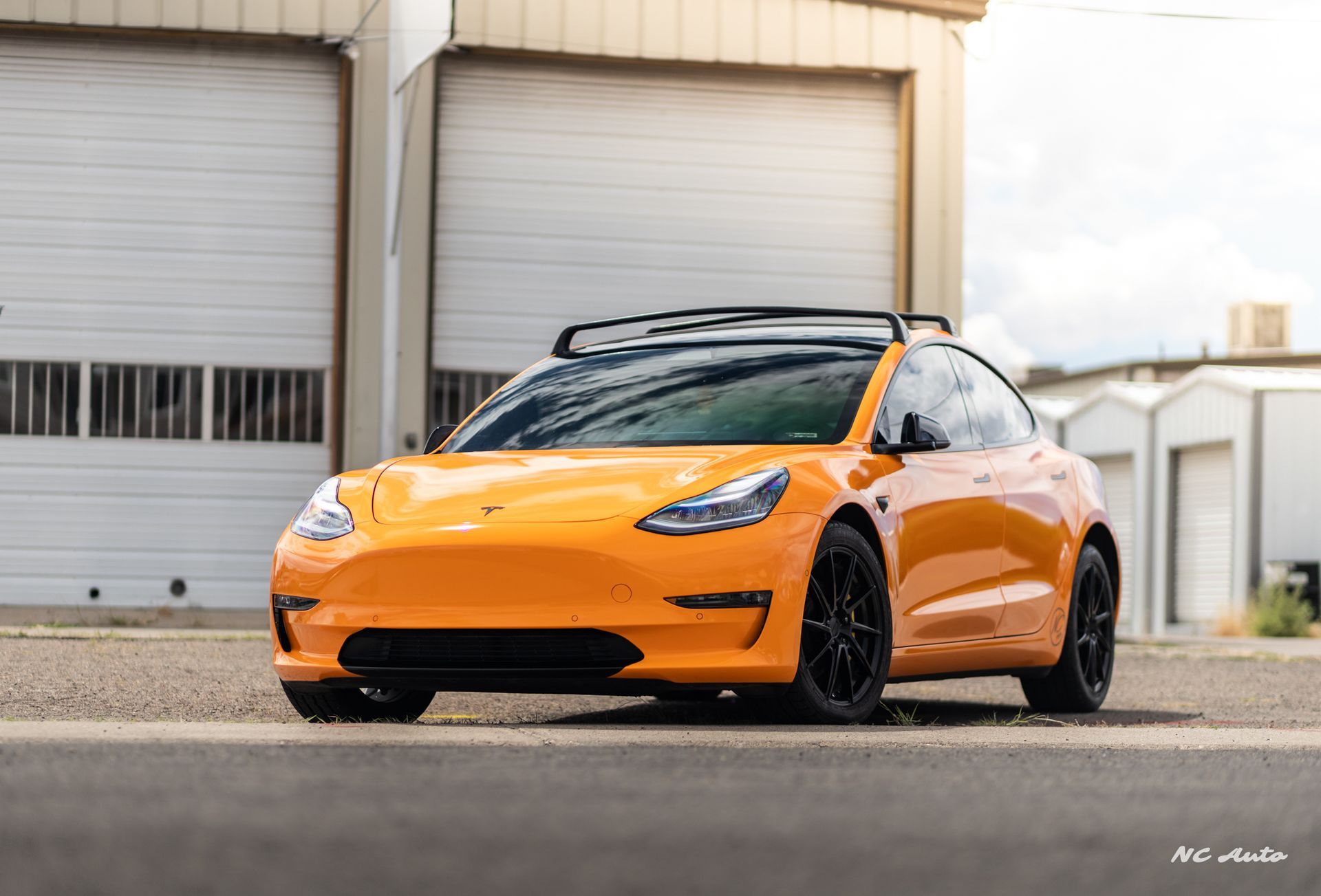 Orange Tesla Model 3 with black roof rack and wheels parked in front of closed garage doors.