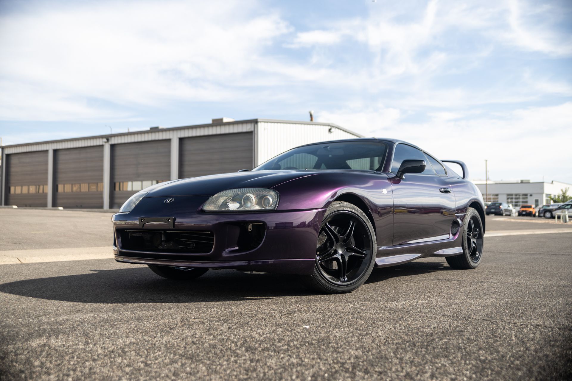 Purple Toyota Supra sports car parked on asphalt. Black wheels, building in background, cloudy sky.
