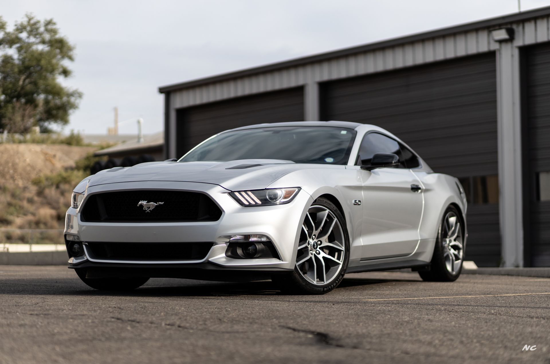 Silver Ford Mustang parked in front of a garage, with gray wheels and a bright sunny day.