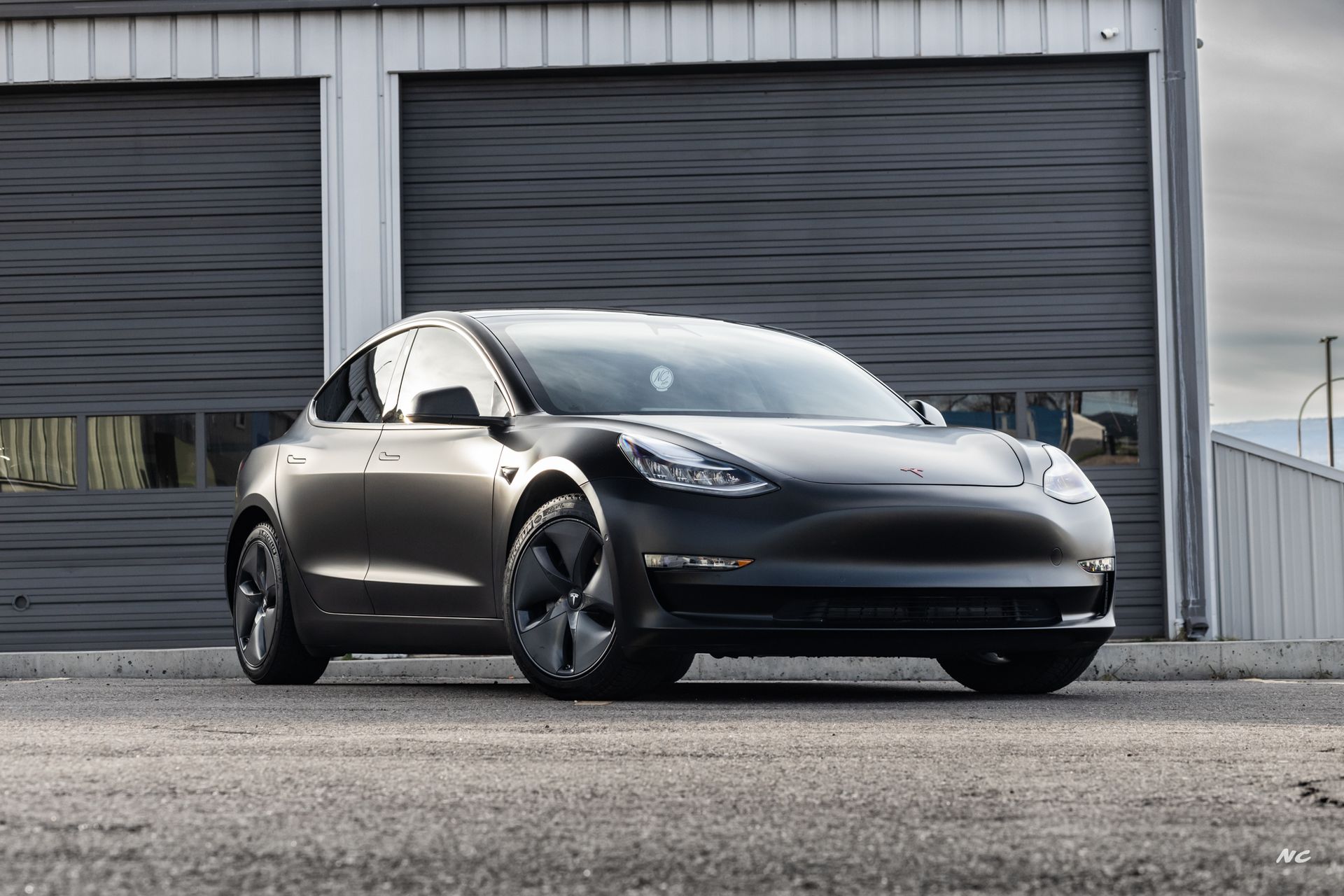 Dark gray Tesla Model 3 electric car parked in front of a gray garage door.