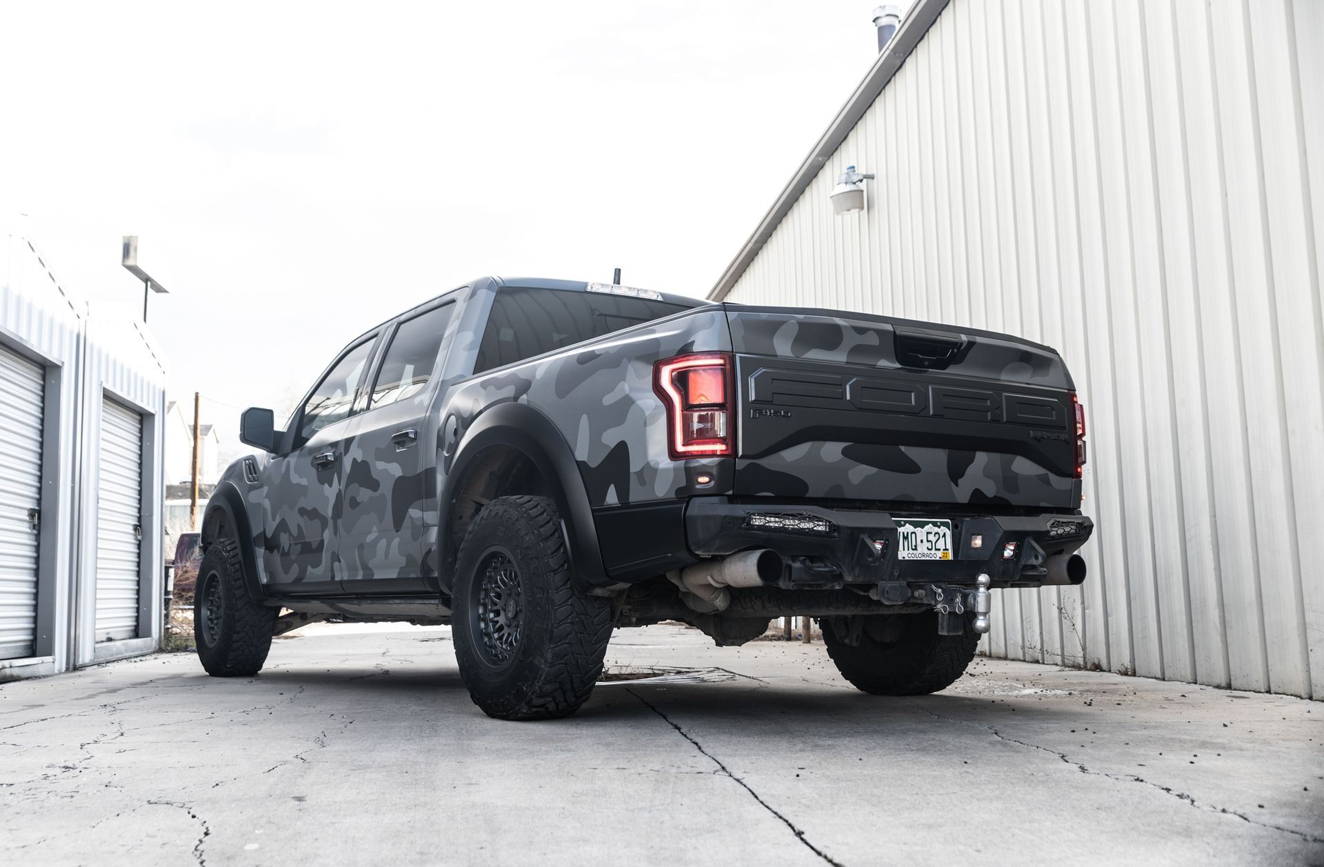 Gray camouflage Ford Raptor pickup truck parked in an alley beside a white building.