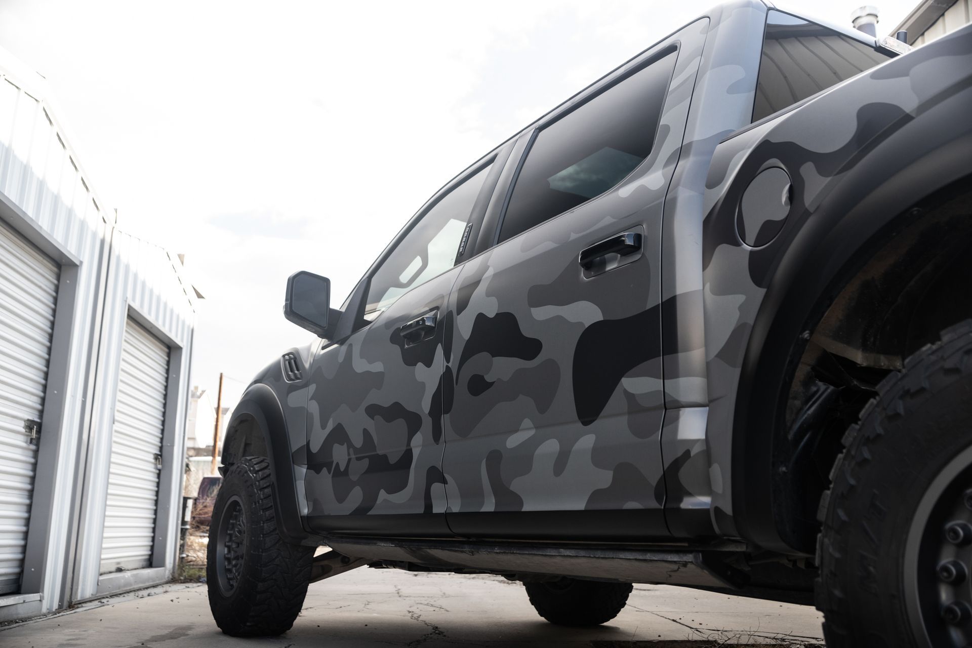 Gray camouflage Ford Raptor truck parked next to a white building.