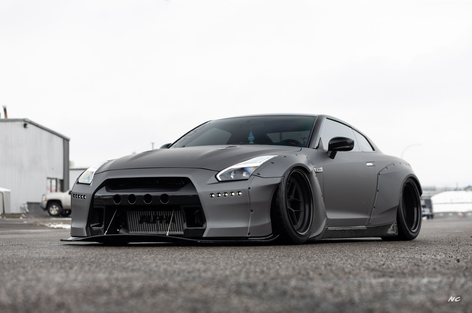 Gray Nissan GT-R sports car with wide body kit, lowered suspension, and black wheels on asphalt.