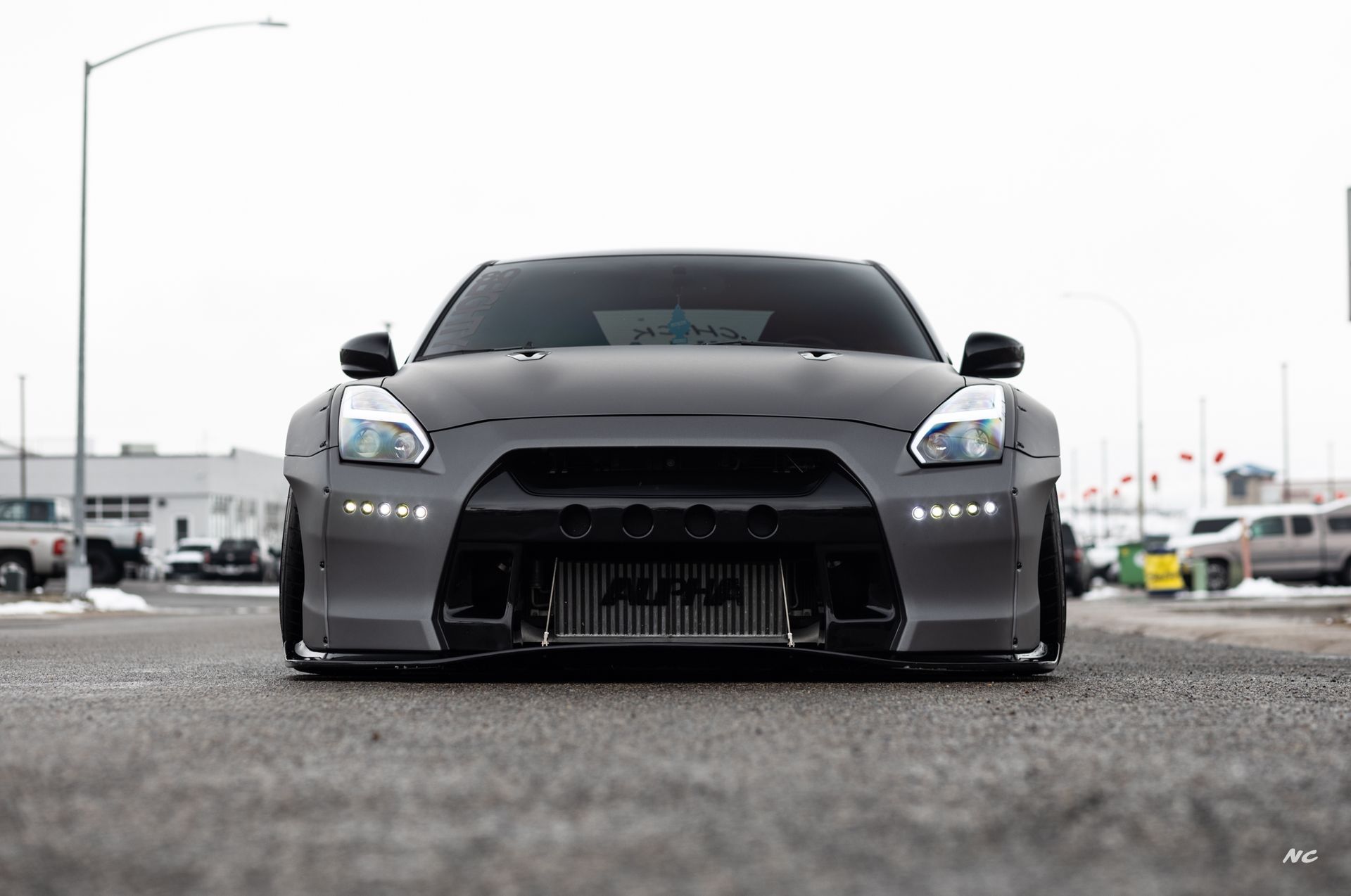 Gray Nissan GT-R sports car with modified front bumper, sitting low on a road. Cloudy, outdoor setting.