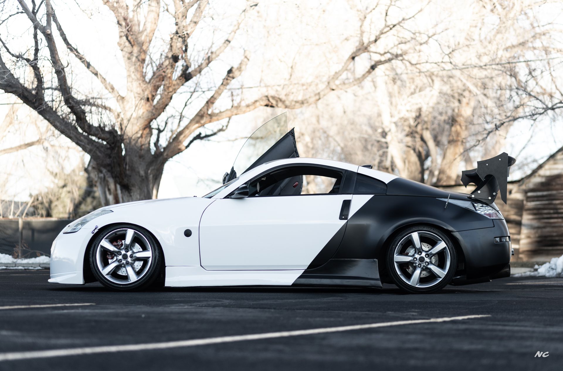 White and black sports car with door open, parked in front of a tree and building.