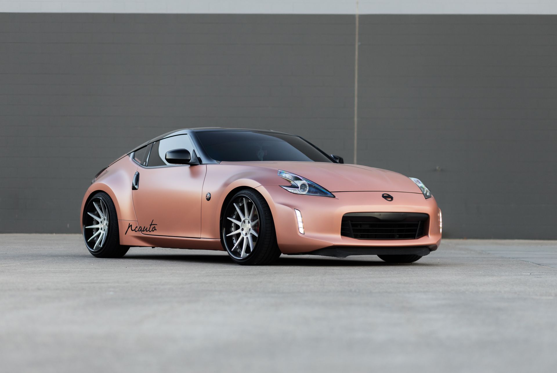 Rose gold Nissan 370Z sports car on a concrete surface against a gray wall.