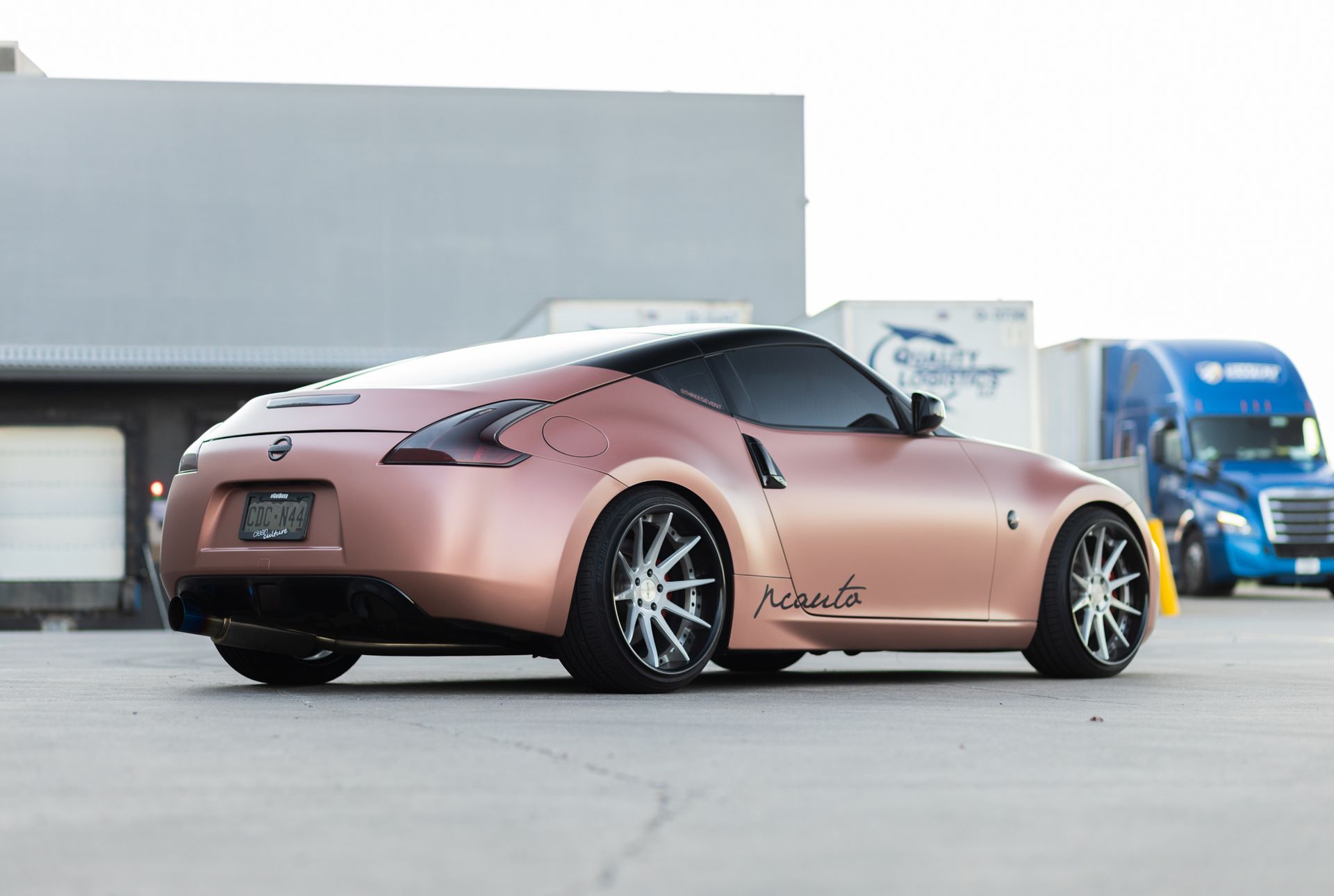 Rose gold Nissan 370Z sports car, low profile, custom wheels, in front of a building and truck.
