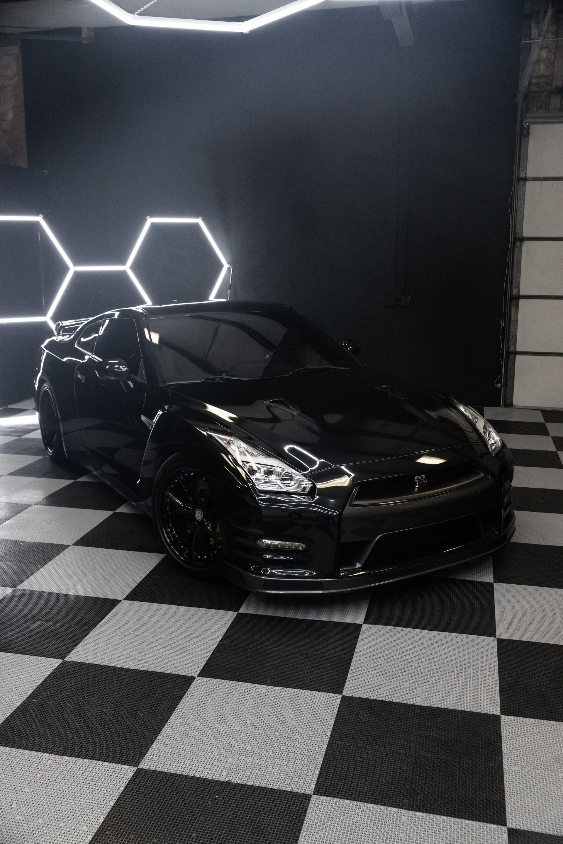 Black Nissan GT-R sports car parked on checkered floor in garage under hexagonal lights.