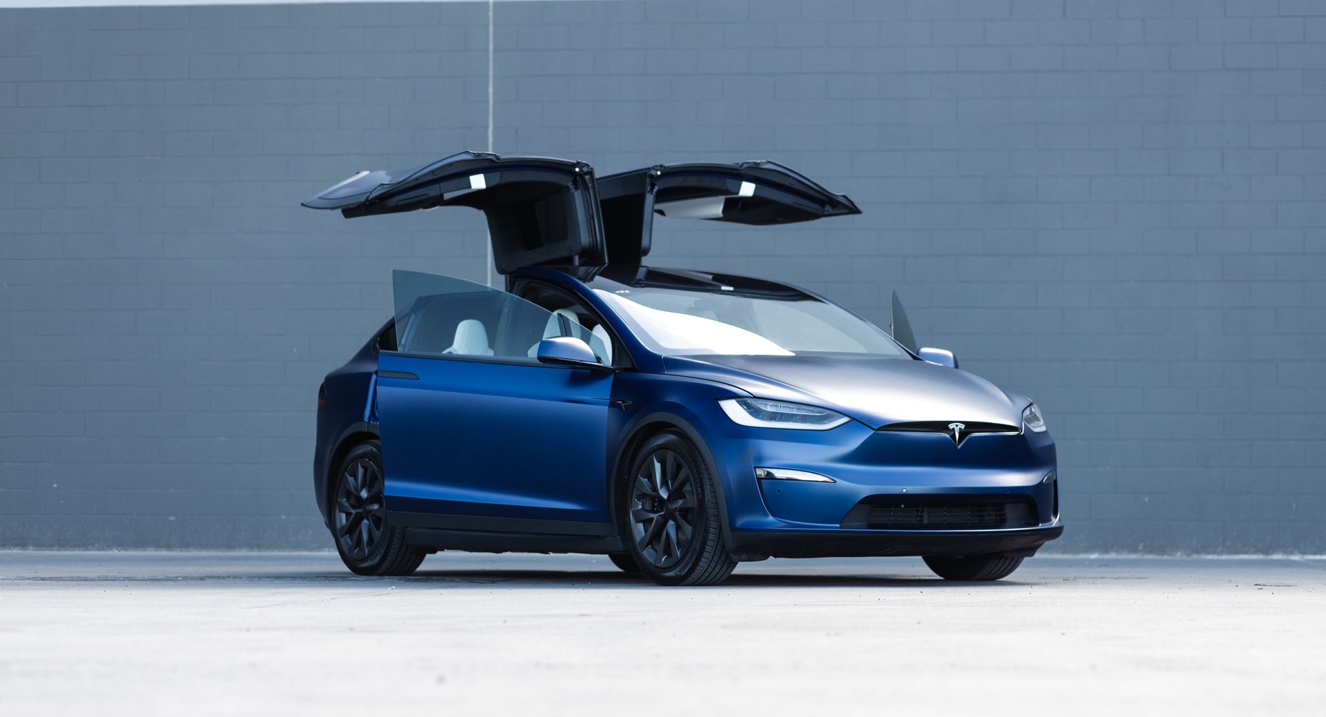 Blue Tesla Model X with falcon-wing doors open, parked in front of a gray wall.