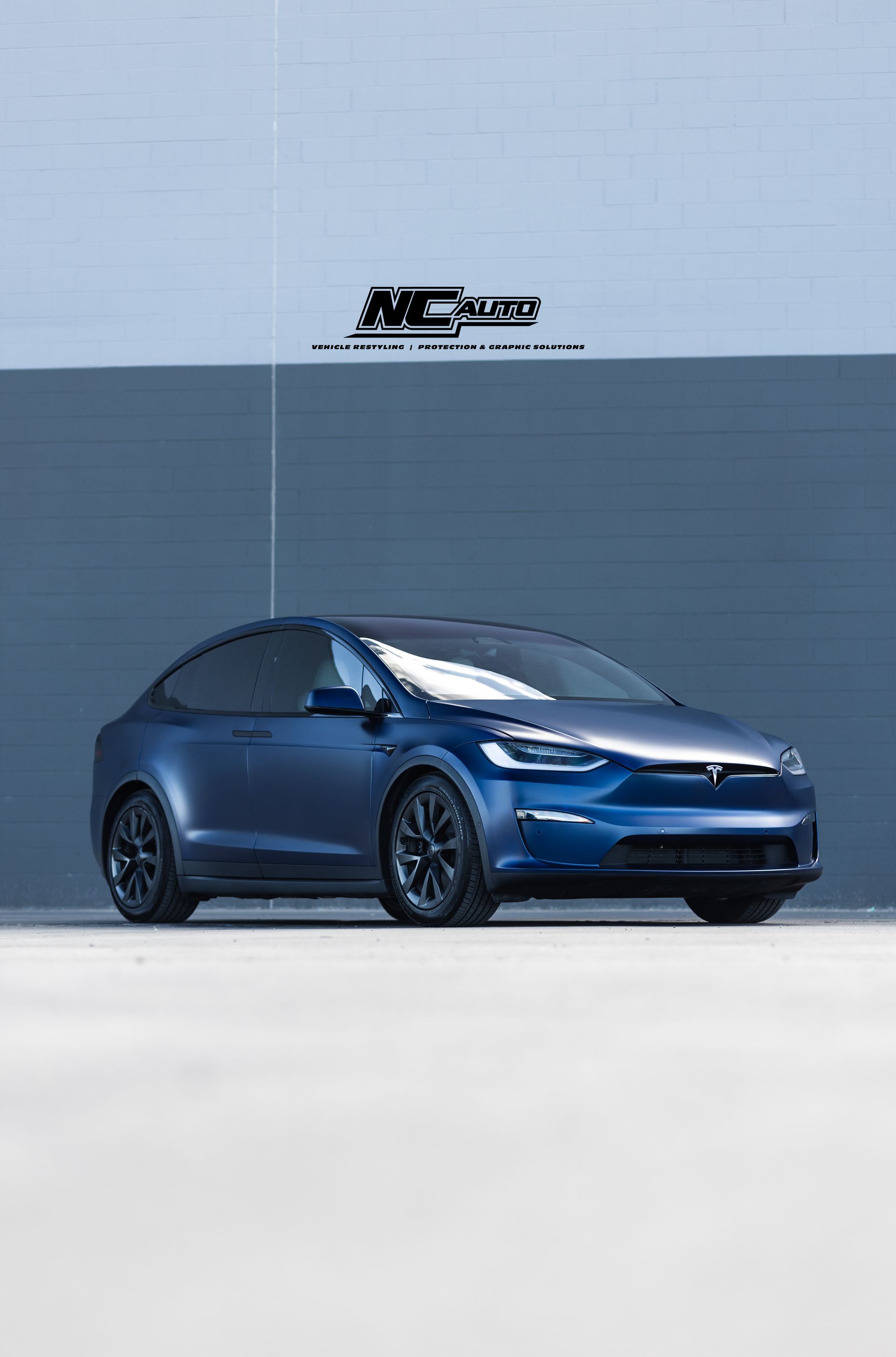 Blue Tesla Model X parked in front of a blue wall. Black wheels.