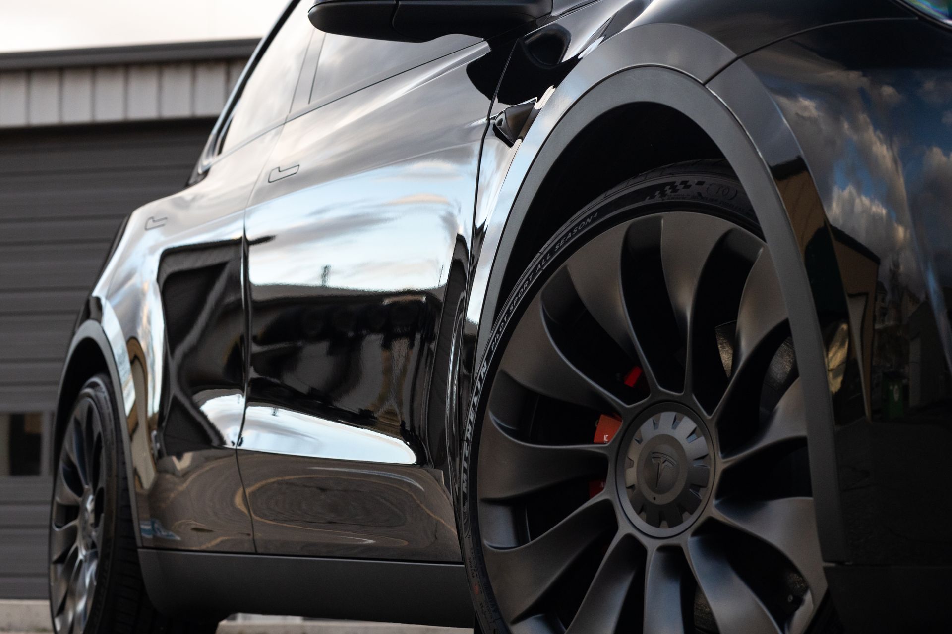 Black Tesla Model Y, glossy paint, turbine-style wheel, red brake caliper, garage door in background.