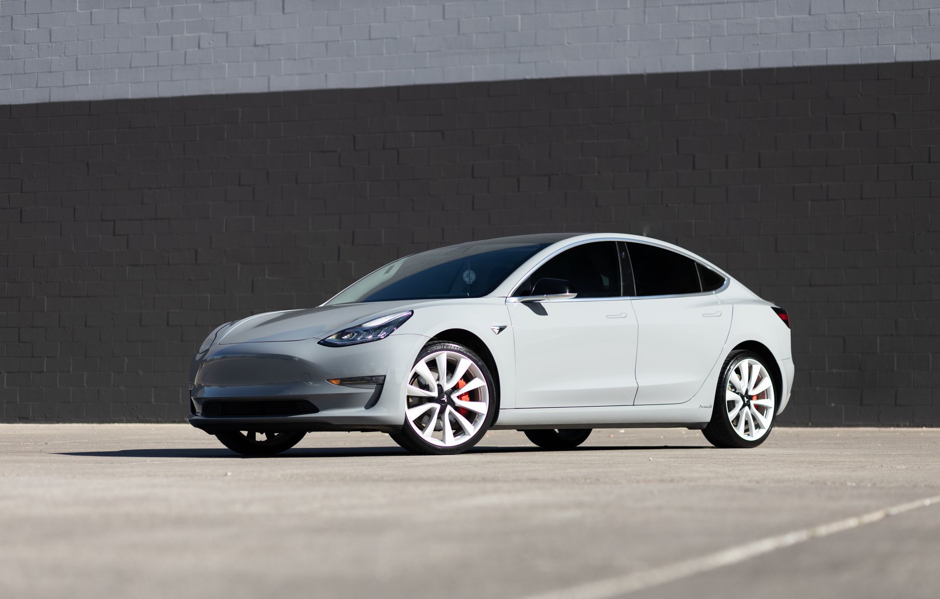 Gray Tesla Model 3 car parked in front of a gray wall, sunny day.