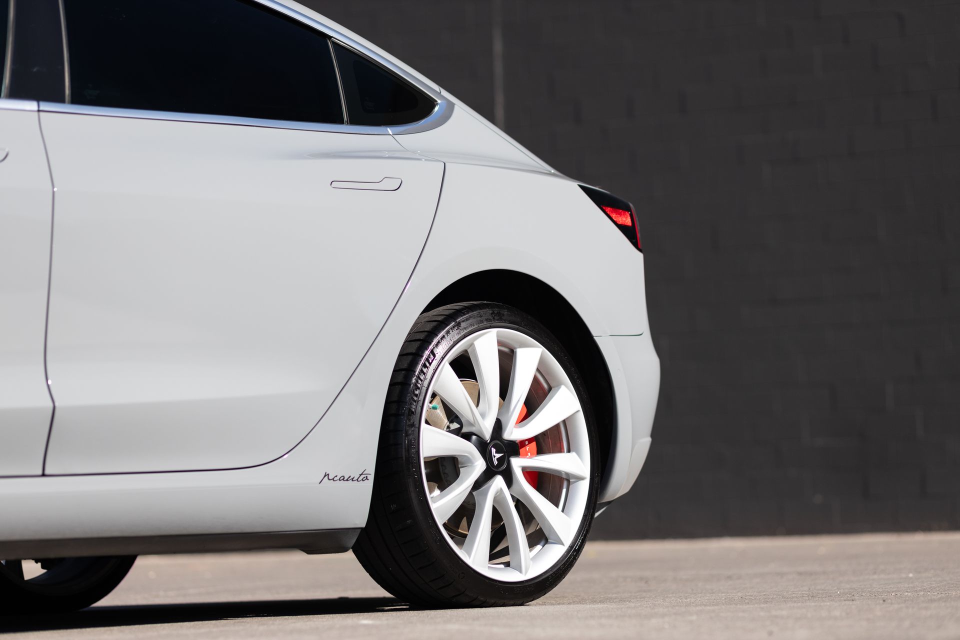 White Tesla Model 3, rear view, white wheels, red brake calipers, against a gray wall.