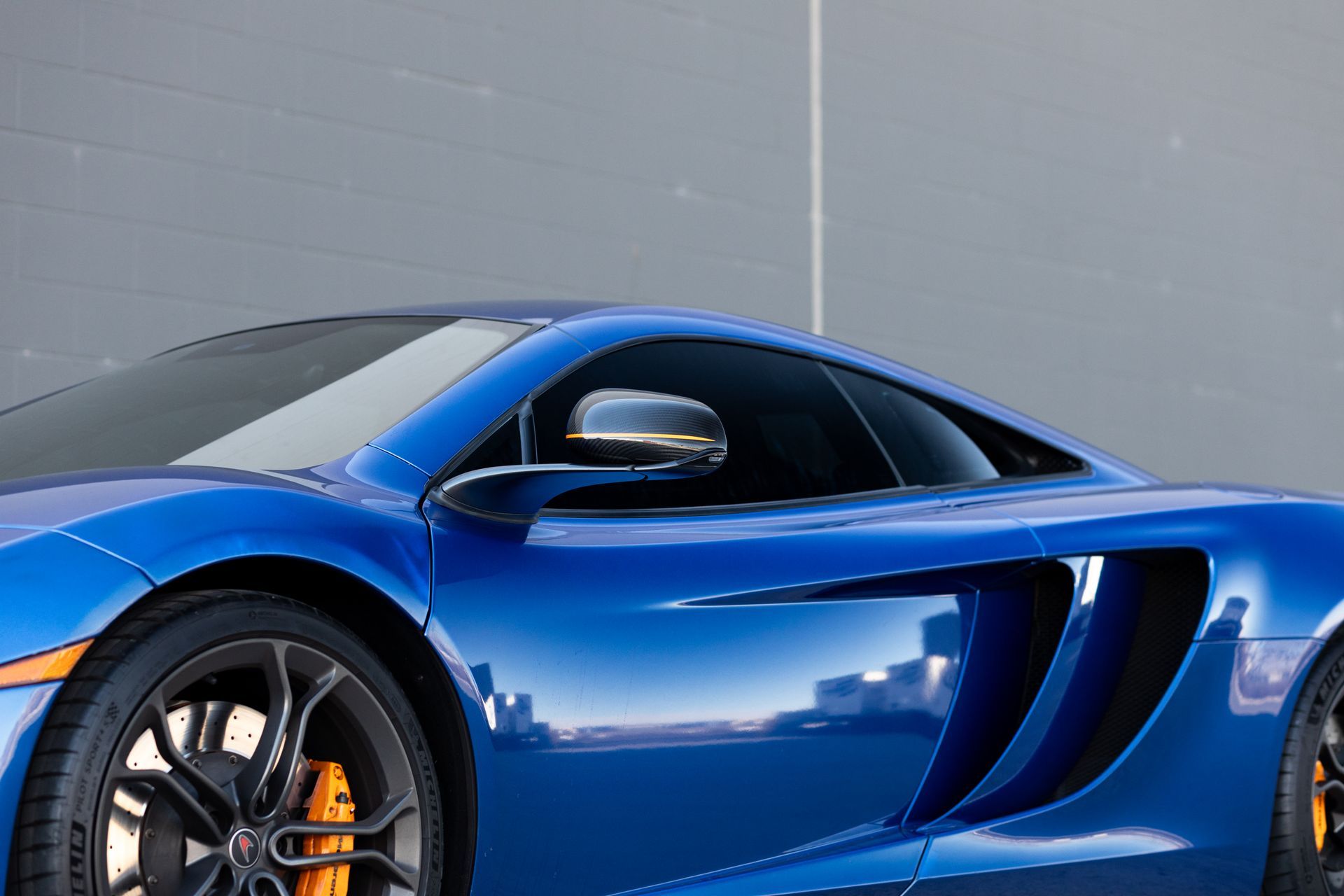 Blue sports car with black wheels and orange brake calipers parked in front of a gray wall.