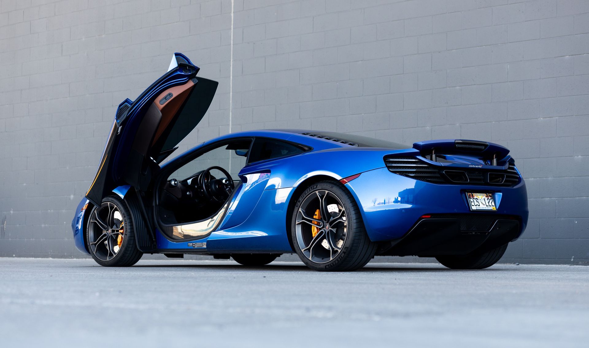 Blue McLaren sports car with doors open, parked against a gray wall.