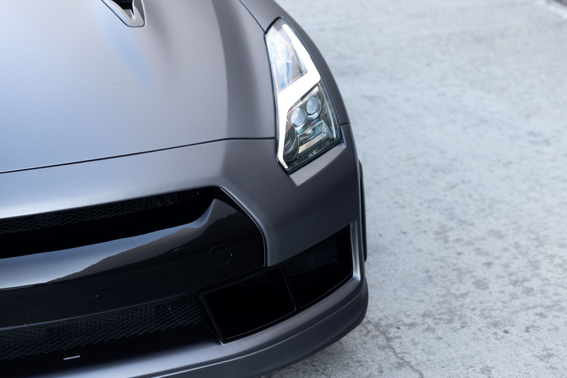 Gray Nissan GT-R sports car front corner, showing headlight, hood vent, and grille, on a textured concrete surface.