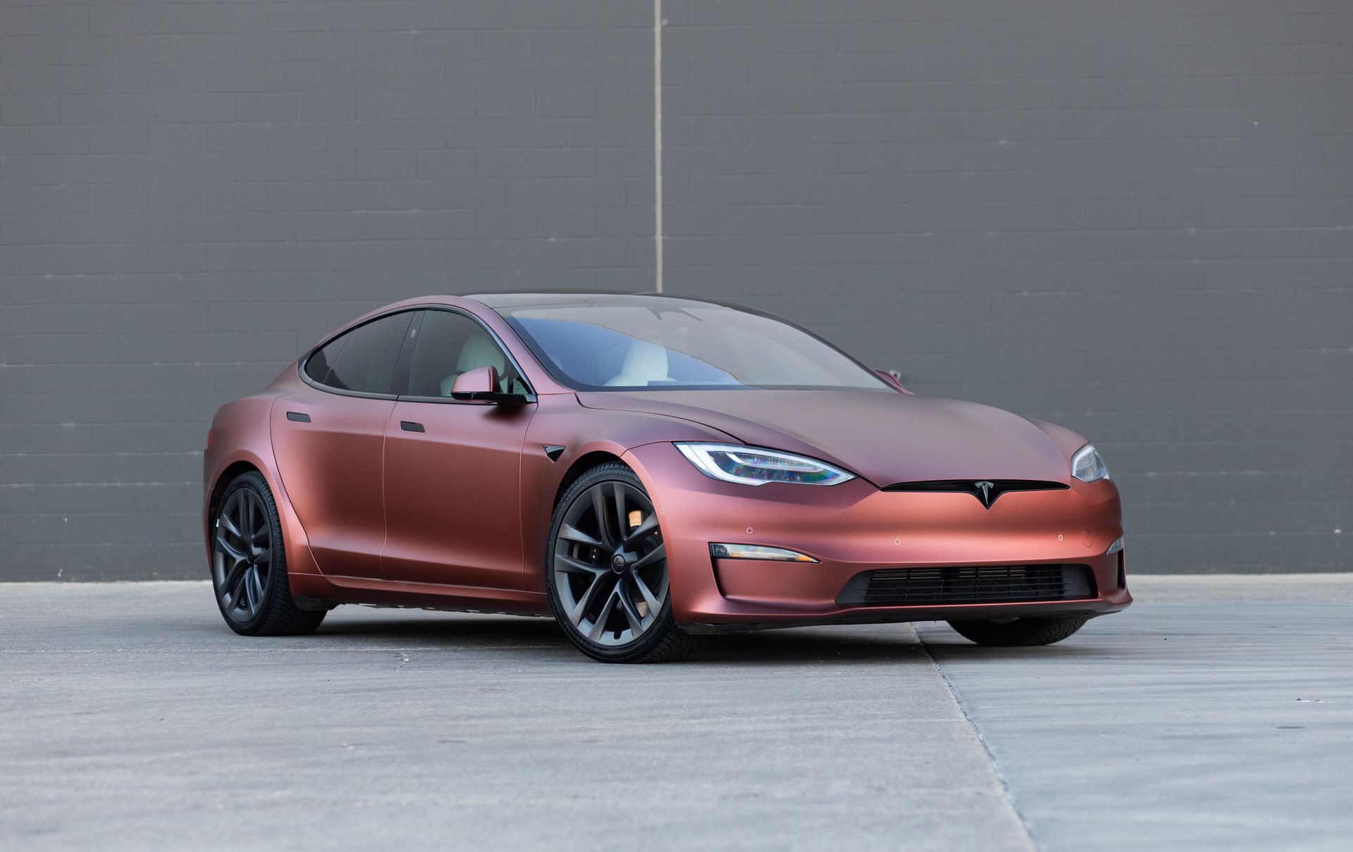 Matte reddish-brown Tesla Model S car parked in front of a gray wall.