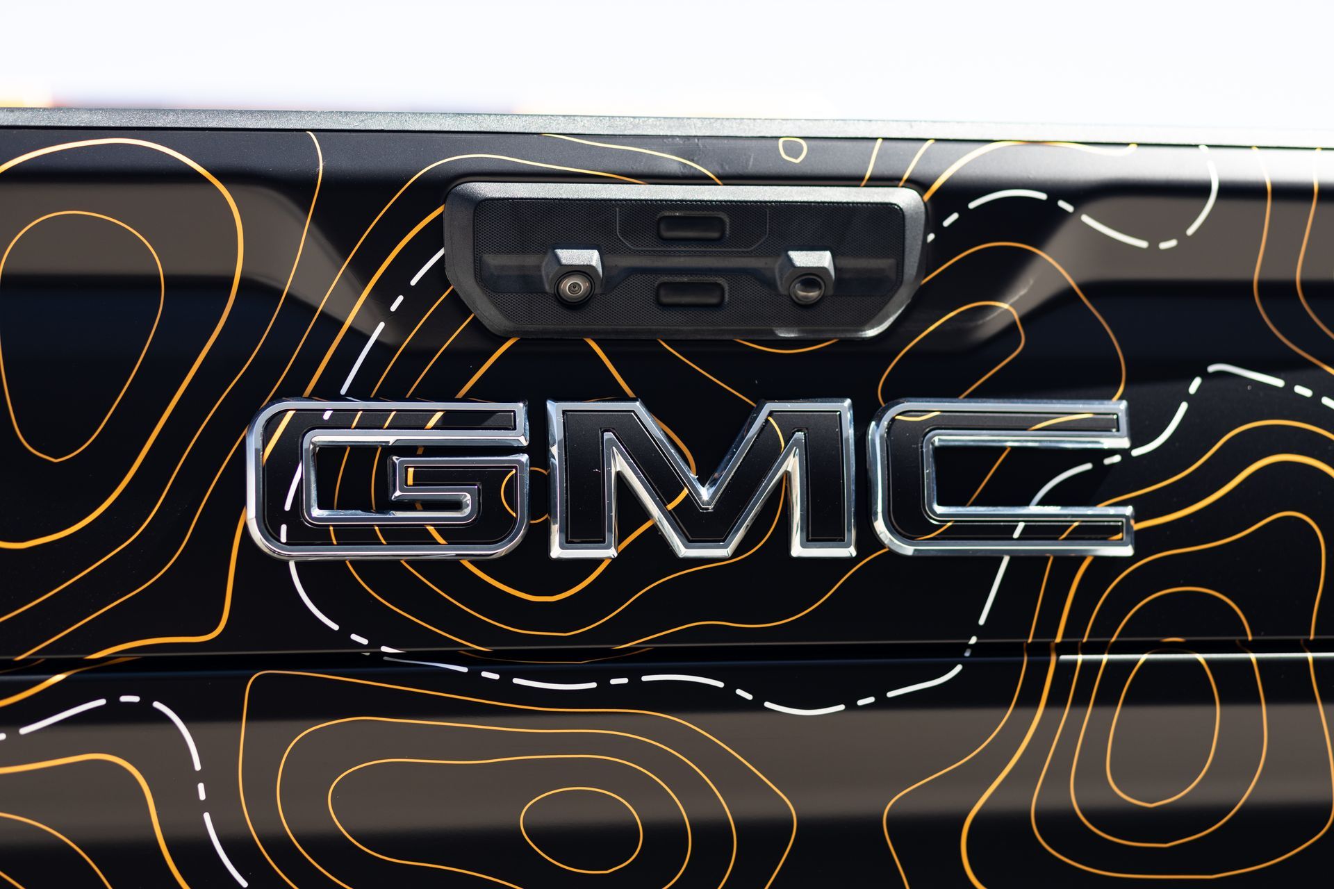 GMC tailgate with chrome lettering and camera, set against a black and gold topographic pattern.