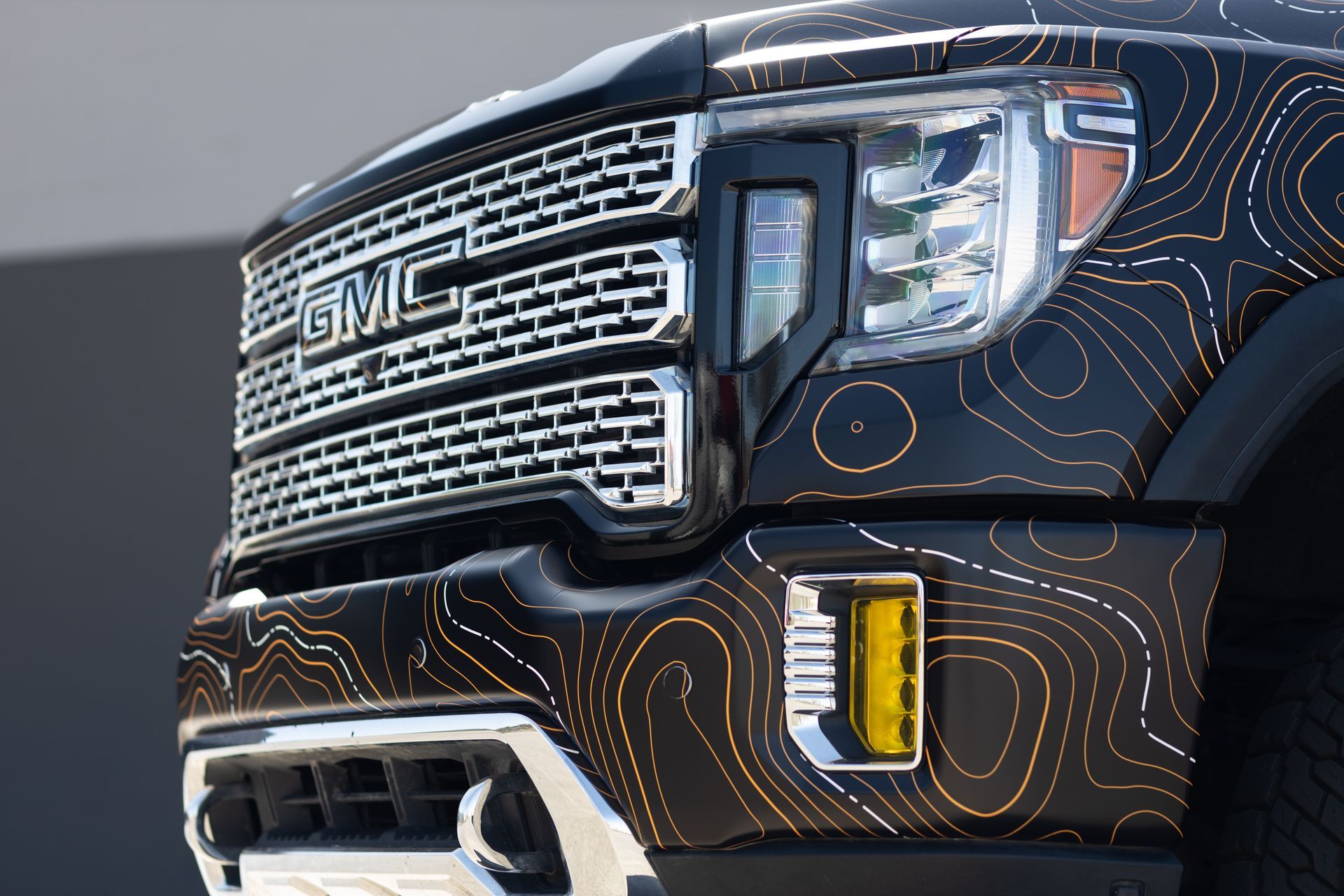 Close-up of a black GMC truck's front with a topographic pattern, a chrome grille, and yellow fog lights.