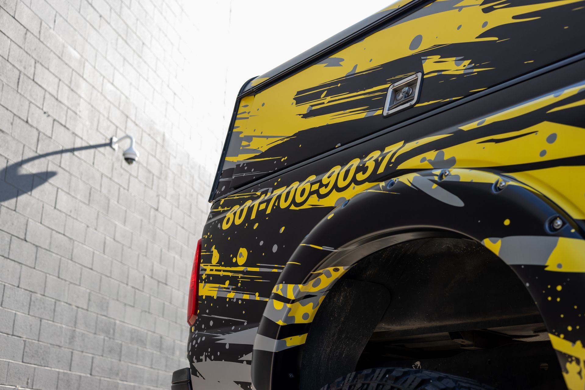 Close-up of a vehicle's back quarter with a black and yellow pattern, near a gray brick wall and a security camera.