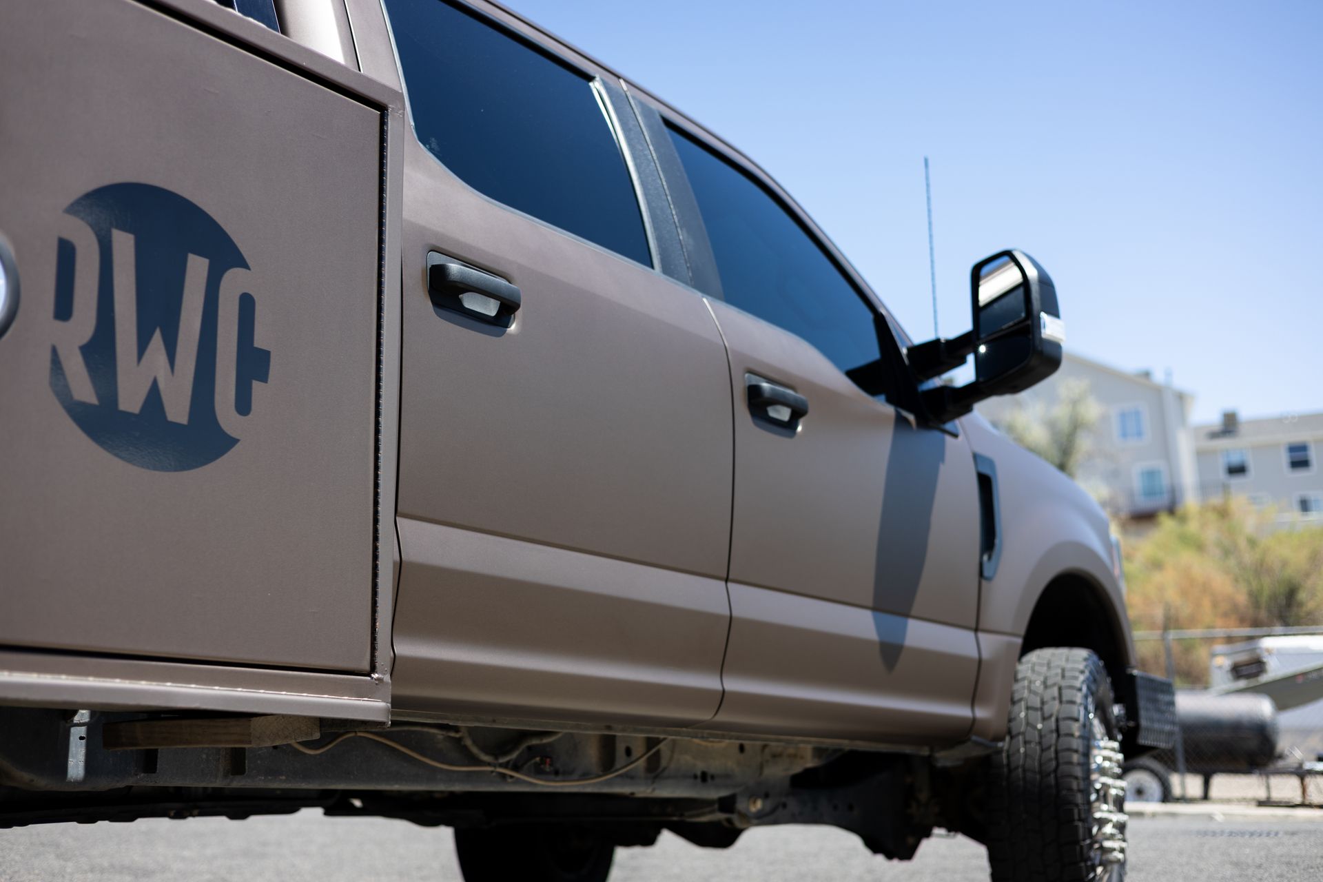 Brown work truck with dark-tinted windows parked outdoors on a sunny day. The side has a circular company logo.
