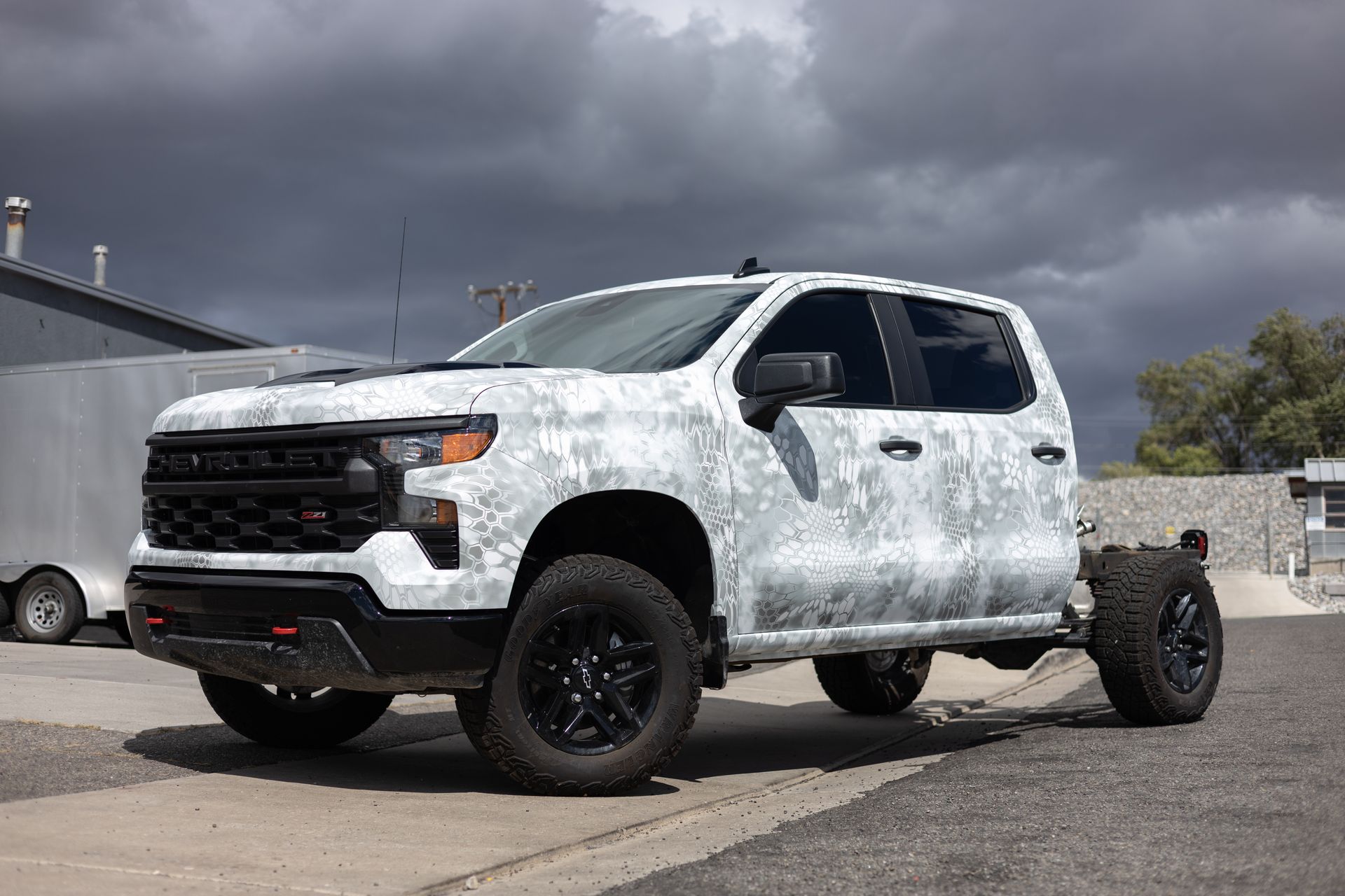 Camouflage-wrapped Chevrolet pickup truck missing its bed, parked outside, under cloudy sky.
