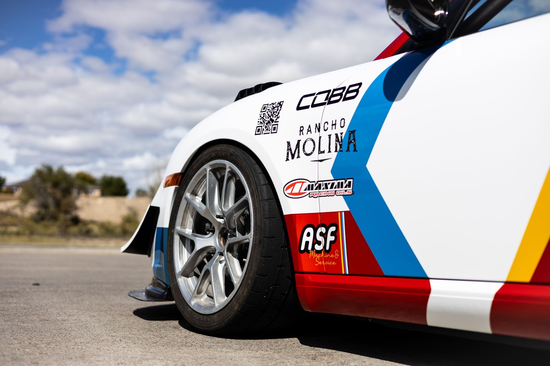 White and red race car side with sponsor logos, silver wheel, and a blue sky in the background.