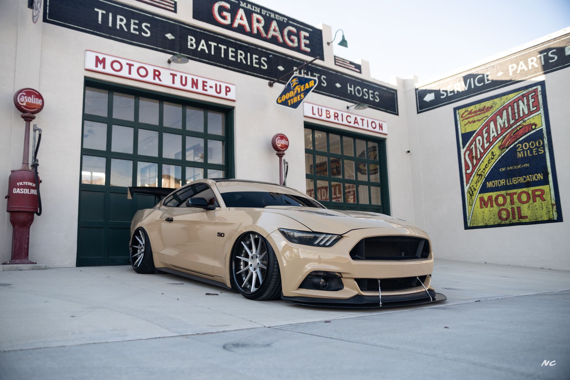 Tan Ford Mustang lowered in front of a vintage garage with gas pumps and signs.