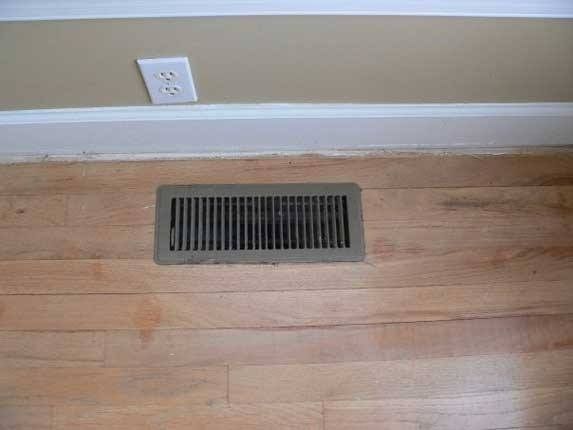 Floor vent on hardwood floor, beige wall and electrical outlet above.