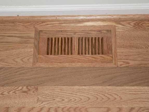 Wooden floor with a rectangular air vent, set against a white baseboard.