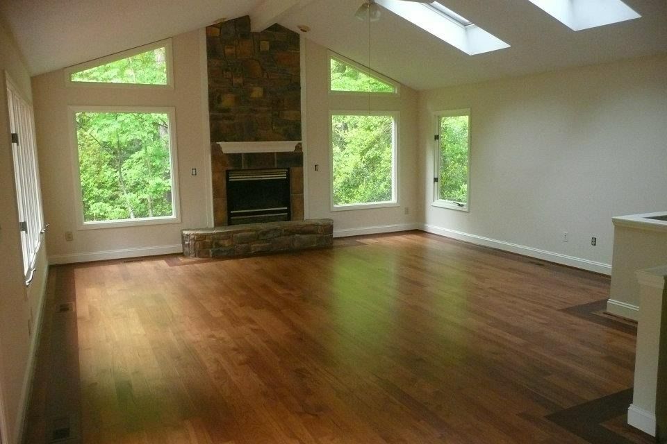 Spacious living room with hardwood floors, stone fireplace, large windows overlooking trees, and skylights.