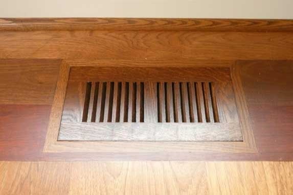 Wooden air vent set into a wooden floor, surrounded by wood trim.