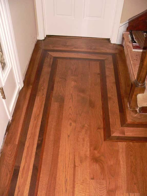 Hardwood floor entryway with dark border, stairs on right, white door in background.