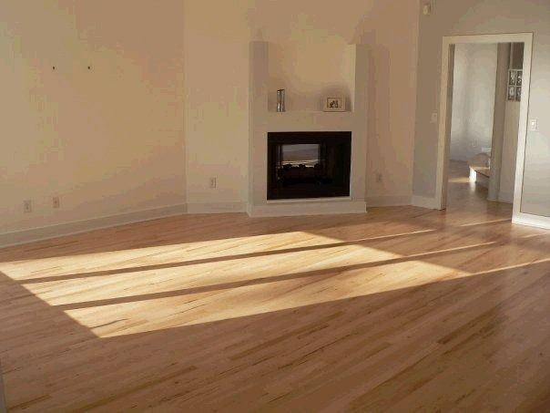 Empty living room with light wood floors, fireplace, and a doorway. Sunlight streams in.