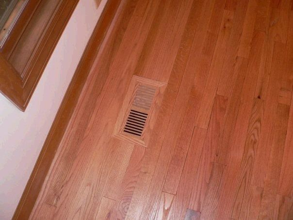 Wooden floor with a vent near a light-colored wall and wooden trim.
