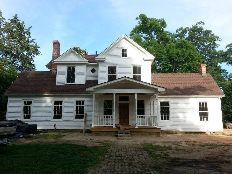White two-story house with brown roof, small porch, windows, and brick walkway. Trees in background.