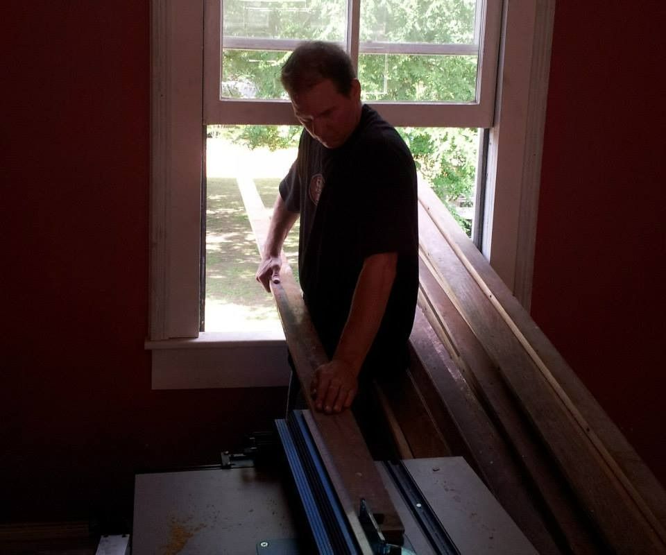Man using a table saw near a window; sawing wood inside a room with red walls.