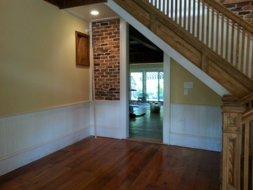 Hallway with wood flooring, brick accent, wainscoting, and wooden staircase. Doorway leads to a bright room.