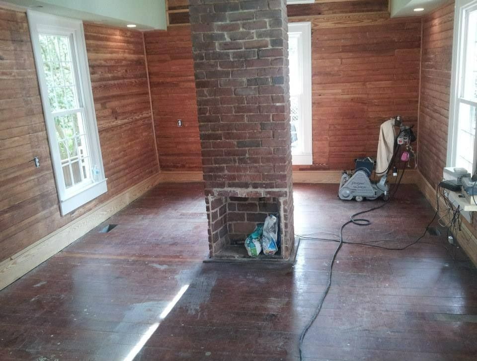 Room interior with exposed brick chimney, wooden walls and floor, two windows.