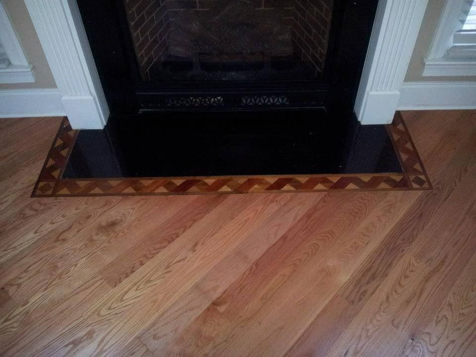 Fireplace with black mantel, stone hearth, and chevron-patterned wood border. Angled wood floor.