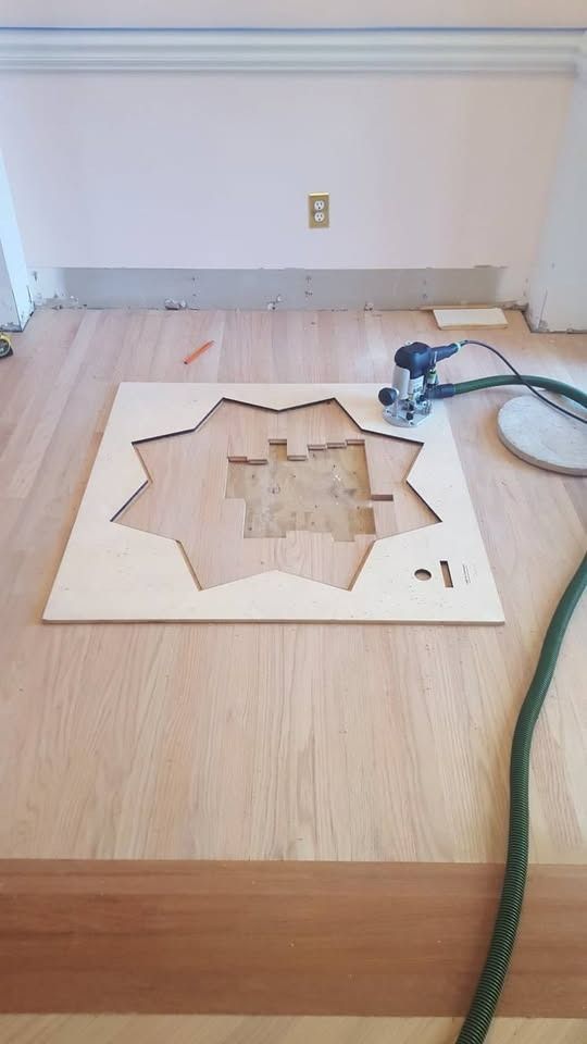 A router cutting a pattern in a wood floor, using a stencil. Tools and dust collection hose are present.