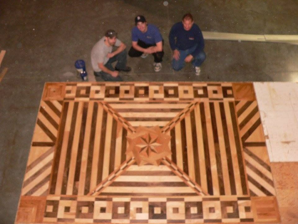 Three men kneel around a complex wood floor inlay with geometric patterns.