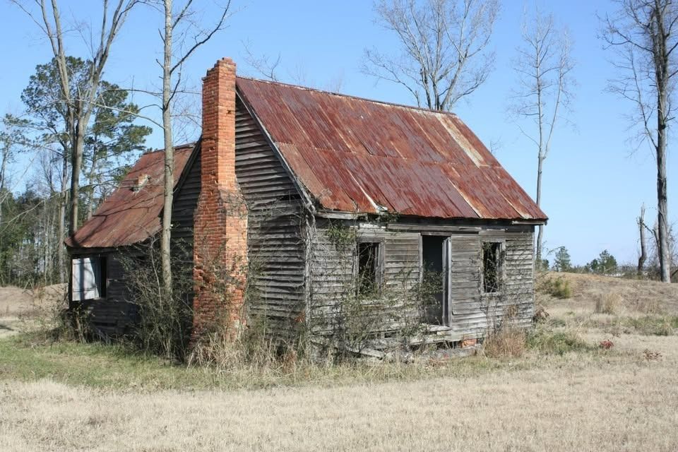Weathered wooden shack with rusty tin roof and brick chimney in a field.