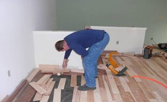 Man installing hardwood floor, kneeling on the floor in a room with green and white walls.