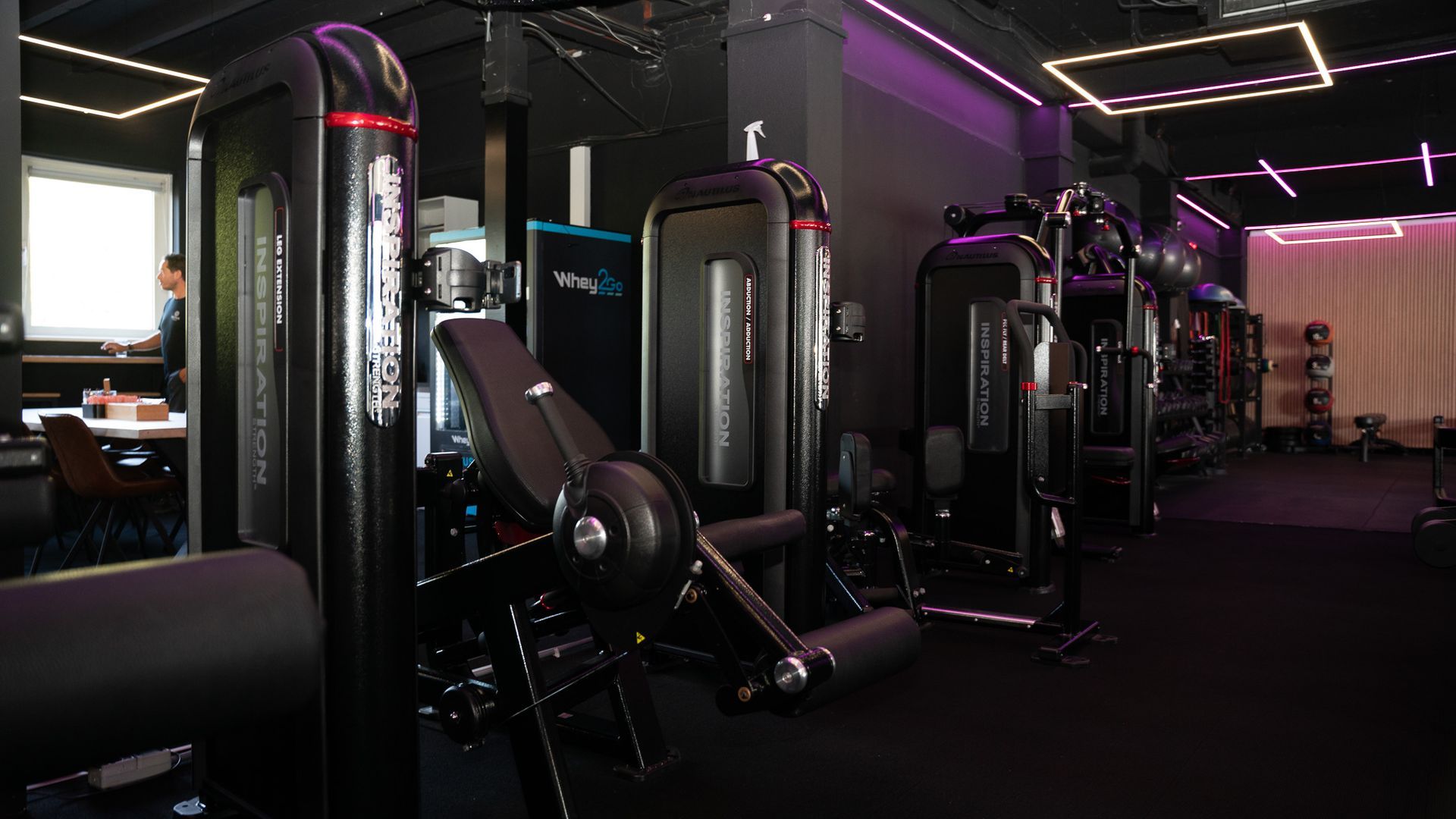 Een sportschool vol met veel fitnessapparatuur en een vrouw op de achtergrond.